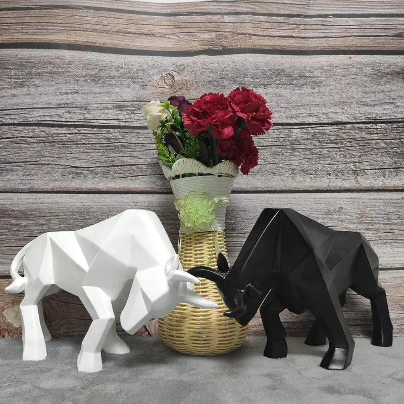 Two geometric bull sculptures, one white and one black, standing next to a vase with flowers on a wooden surface.