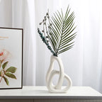 Decorative white vase with green leaves on a white surface, framed artwork in the background.