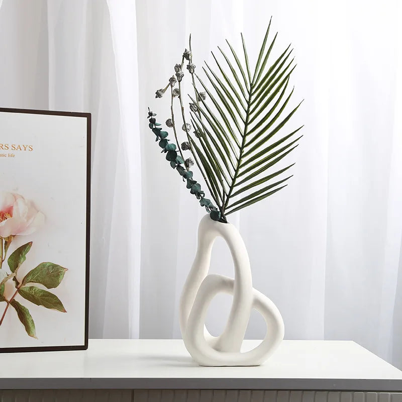 Decorative white vase with green leaves on a white surface, framed artwork in the background.