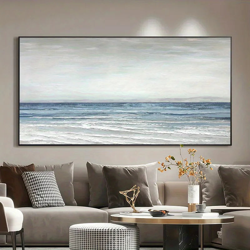 A large horizontal wall art print depicting a calm ocean scene with pale sky and blue-gray waves, displayed above a beige sofa in a modern living room setting.