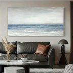 A large horizontal canvas print of a calm blue ocean under a cloudy sky, displayed on a light gray wall above a dark gray leather sofa in a modern living room setting.