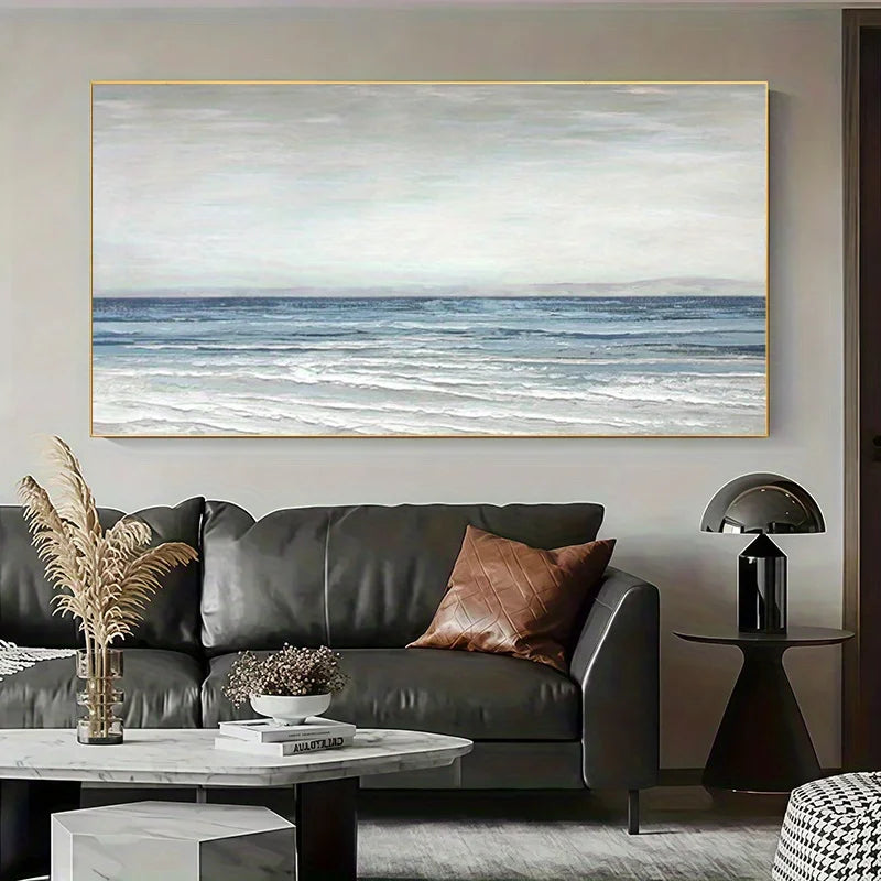 A large horizontal canvas print of a calm blue ocean under a cloudy sky, displayed on a light gray wall above a dark gray leather sofa in a modern living room setting.