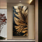 A vertical wall art piece featuring a large metallic gold feather motif on a black background, displayed in a modern interior setting with a beige wall and a window screen to the left.