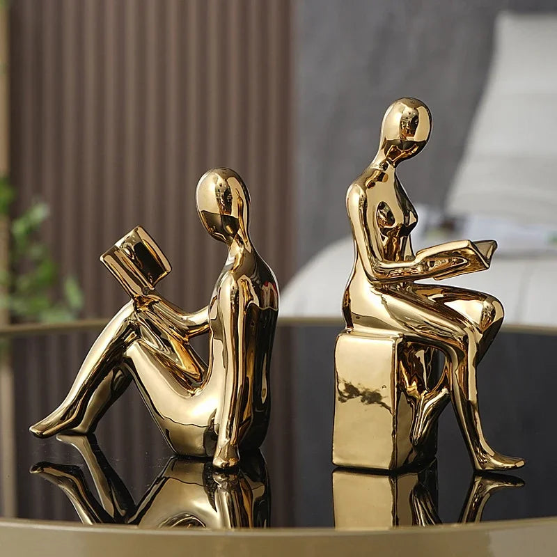 Two gold-colored sculptures of people on a reflective surface with a blurred background