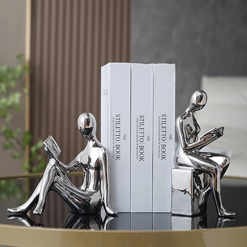Silver human-like bookends holding books on a reflective surface with a blurred background