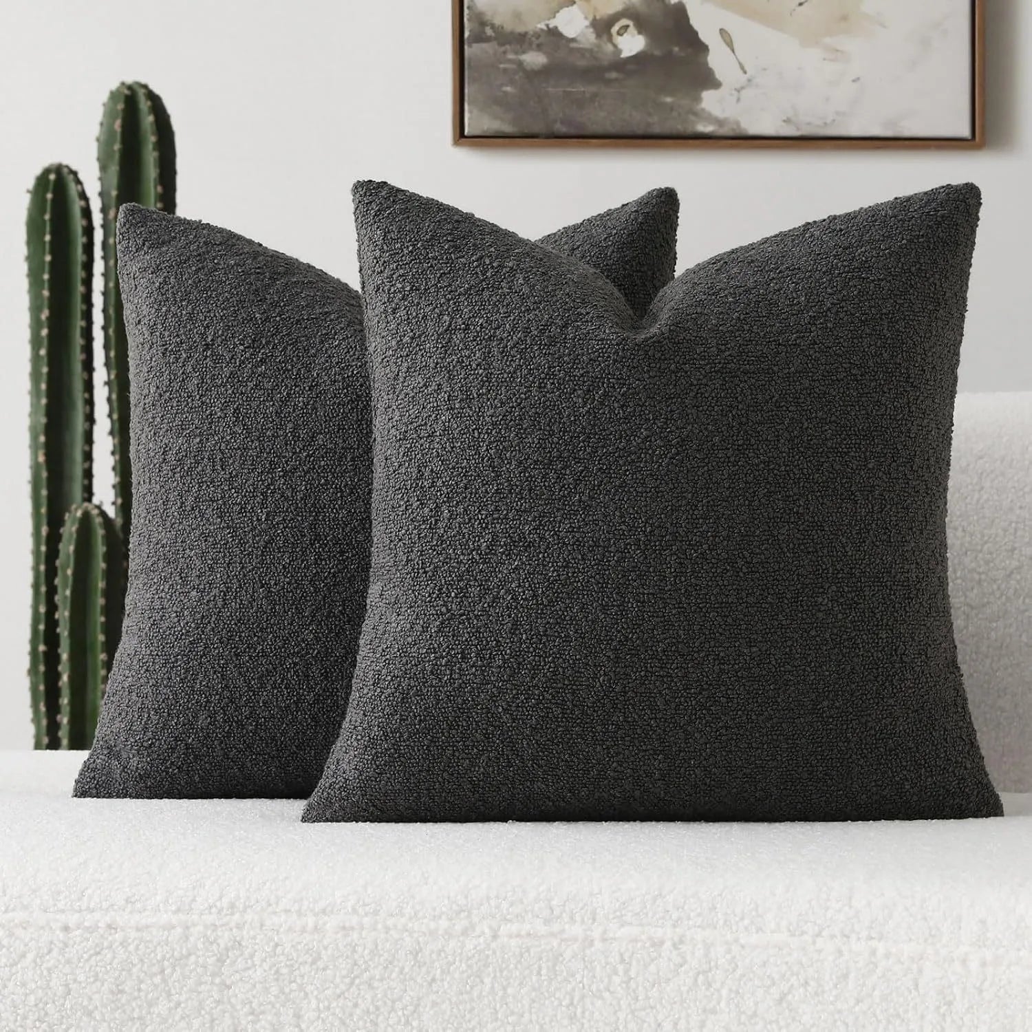 Two dark gray textured pillows on a white sofa with a cactus plant and framed picture in the background.
