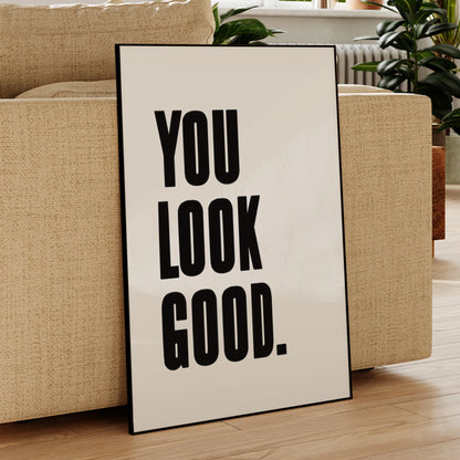 Poster with 'YOU LOOK GOOD.' text leaning against a beige sofa in a living room.