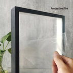 Hand holding a framed picture with protective film on a gray wall background