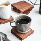 A gray ribbed ceramic coffee mug filled with black coffee sits on a square wooden coaster on a white surface. Another white ribbed mug with tea is visible in the background on a wooden coaster.
