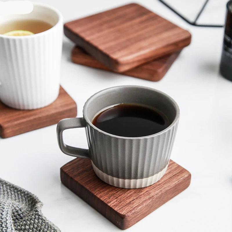A gray ribbed ceramic coffee mug filled with black coffee sits on a square wooden coaster on a white surface. Another white ribbed mug with tea is visible in the background on a wooden coaster.
