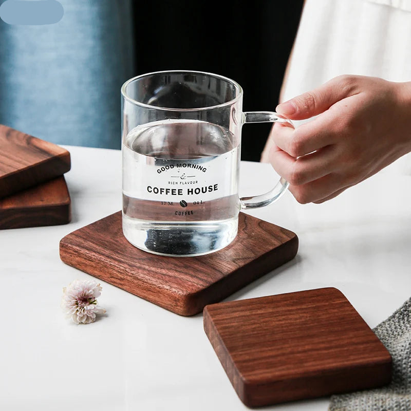 A clear glass mug with a handle sits on a square dark wood coaster, with a hand holding the handle. Another coaster is placed beneath the mug, and a matching coaster lies nearby on a white tabletop.