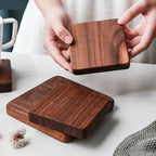 A person holds a square dark wood coaster above a matching stack of two coasters on a light tabletop. The product appears to be wooden coasters with visible wood grain, shown in a clean indoor setting with hands present.