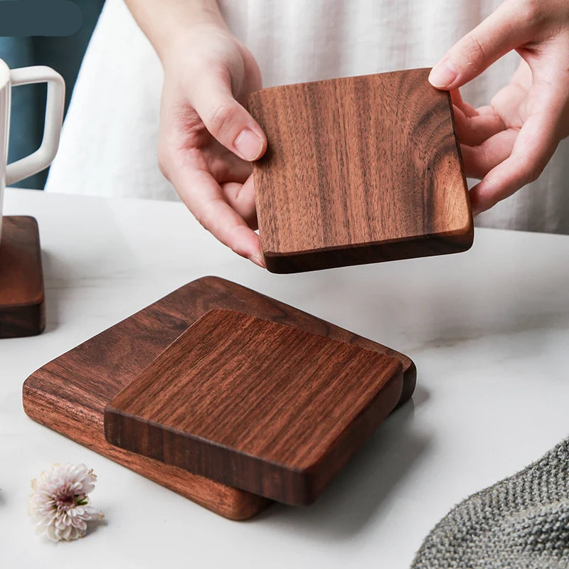 A person holds a square dark wood coaster above a matching stack of two coasters on a light tabletop. The product appears to be wooden coasters with visible wood grain, shown in a clean indoor setting with hands present.