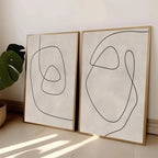 Two abstract line art prints in gold frames on a light surface with a plant in the corner.