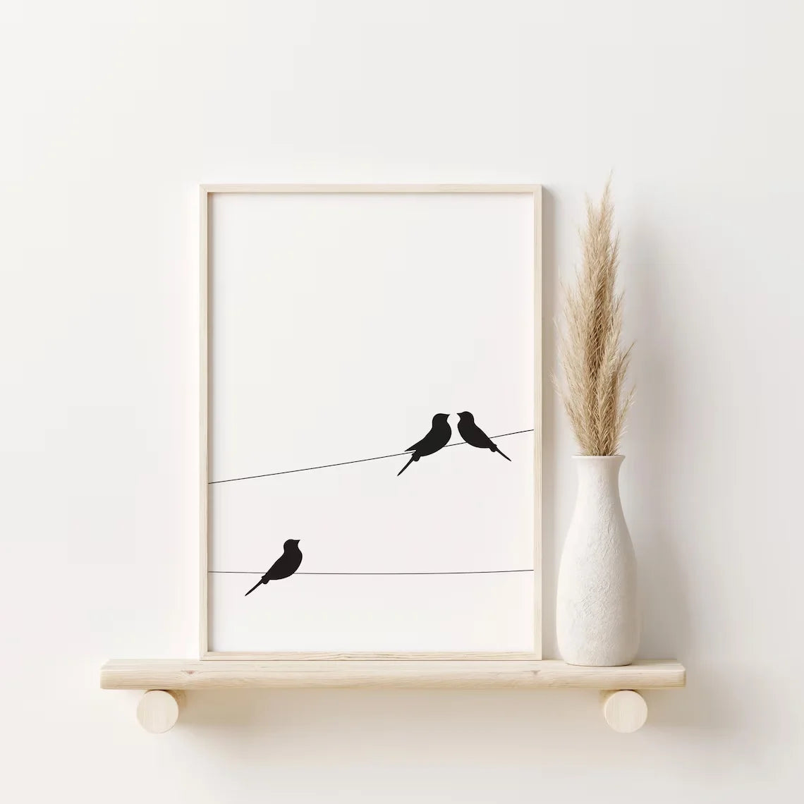 A minimalist wall art print featuring three black bird silhouettes perched on thin horizontal wires, displayed on a light wood floating shelf beside a white textured vase with dried pampas grass against a white wall.