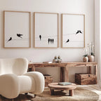 Three wall art prints with black bird silhouettes on thin wires are displayed above a rustic wooden console table in a bright, modern living room. The room features a cream boucle armchair, a round wooden coffee table, and neutral decor.