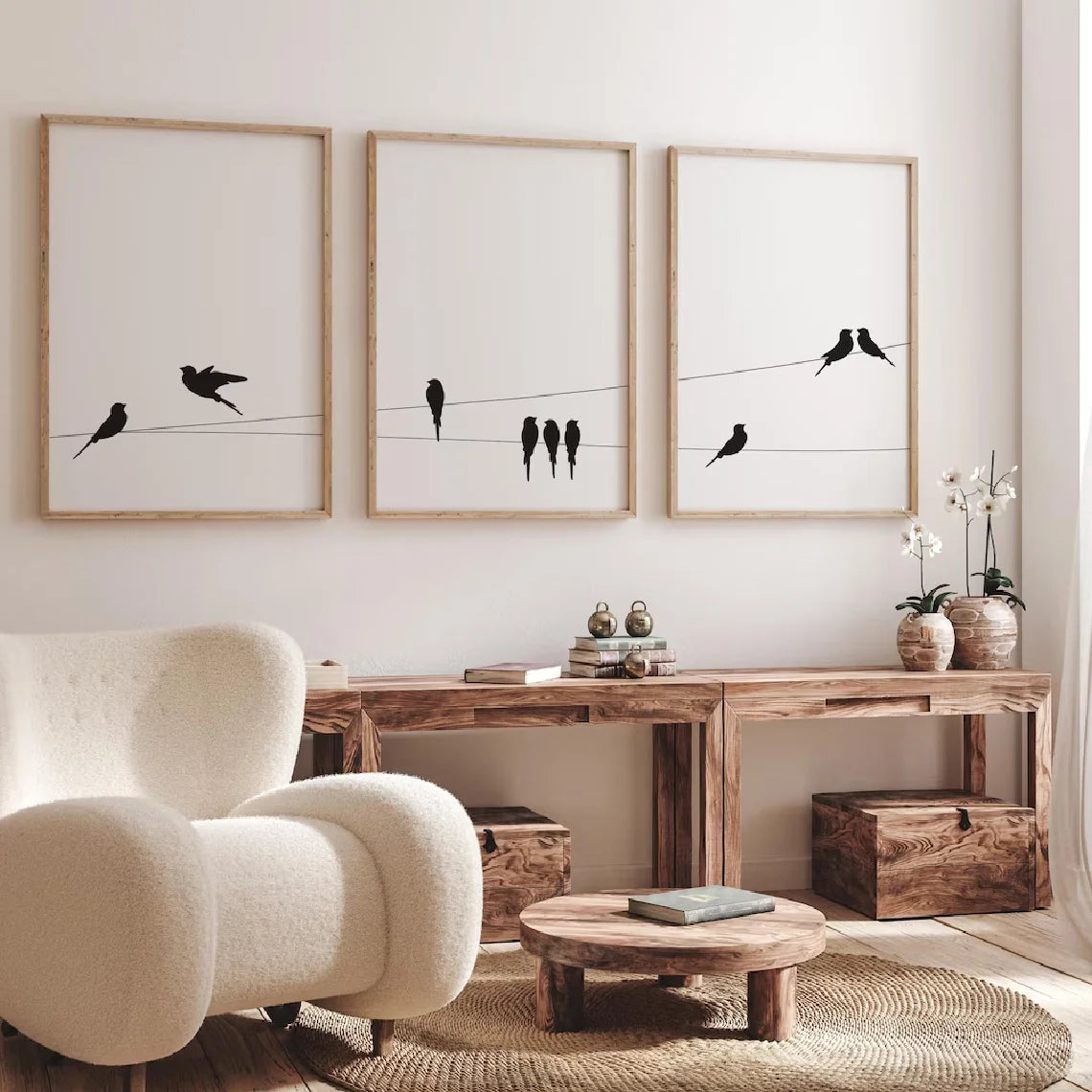 Three wall art prints with black bird silhouettes on thin wires are displayed above a rustic wooden console table in a bright, modern living room. The room features a cream boucle armchair, a round wooden coffee table, and neutral decor.
