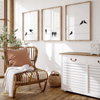 Three framed wall art prints with black bird silhouettes on wires hang above a rattan chair and a white cabinet in a bright, sunlit room with wood floors and a plant.