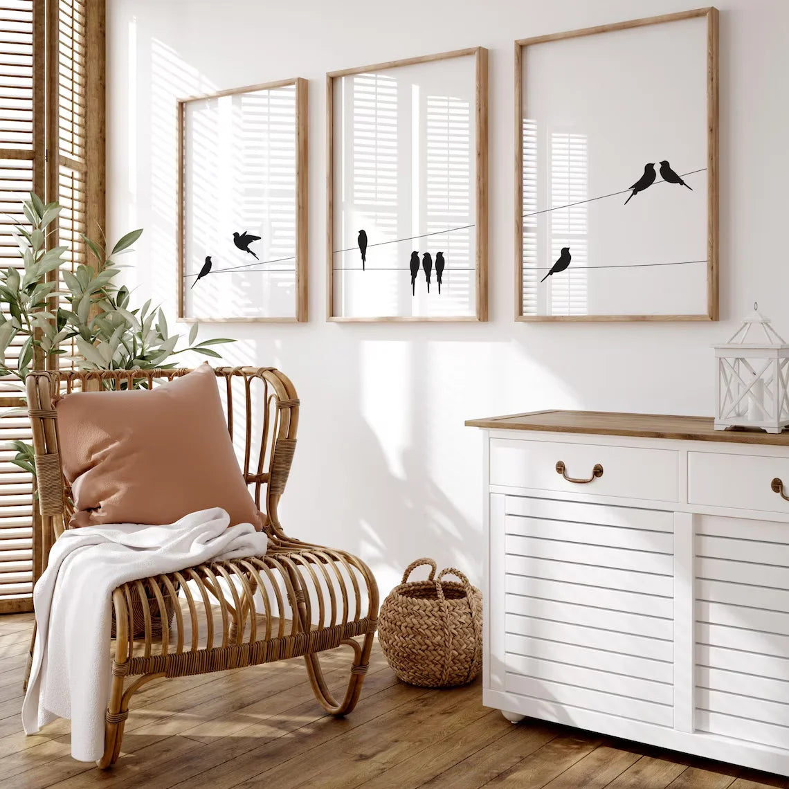 Three framed wall art prints with black bird silhouettes on wires hang above a rattan chair and a white cabinet in a bright, sunlit room with wood floors and a plant.