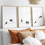 Three framed wall art prints with black bird silhouettes on thin wires, displayed above a bed with white and rust-colored pillows in a bright bedroom.