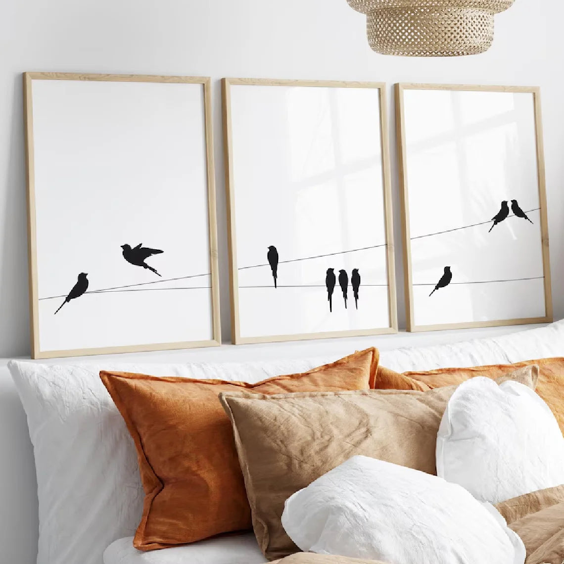 Three framed wall art prints with black bird silhouettes on thin wires, displayed above a bed with white and rust-colored pillows in a bright bedroom.