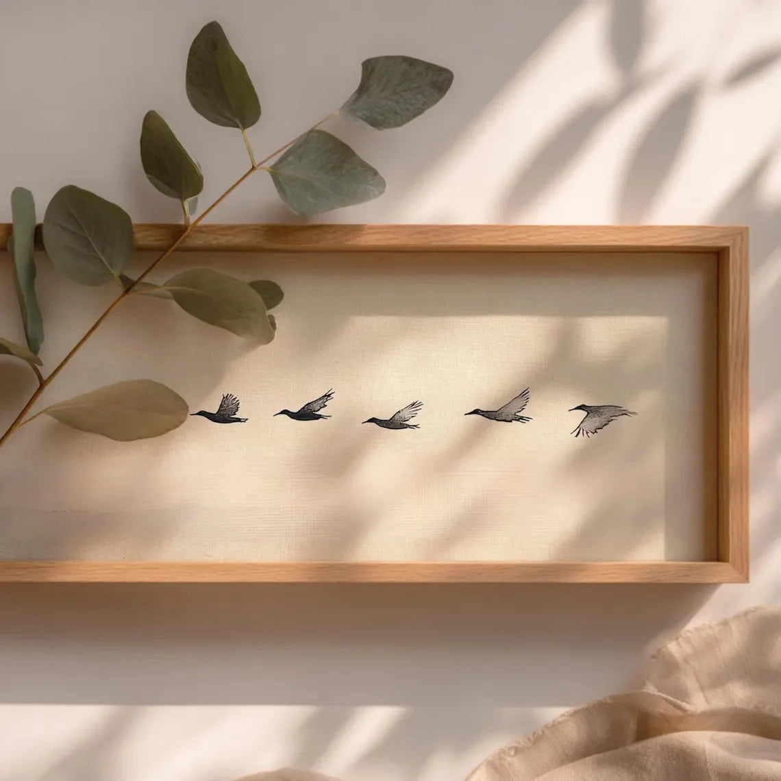 Framed artwork of five birds in flight on a beige background, with a branch of eucalyptus leaves on the left.