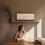 Woman sitting at a table with books and a mug, sunlit room with wall art.