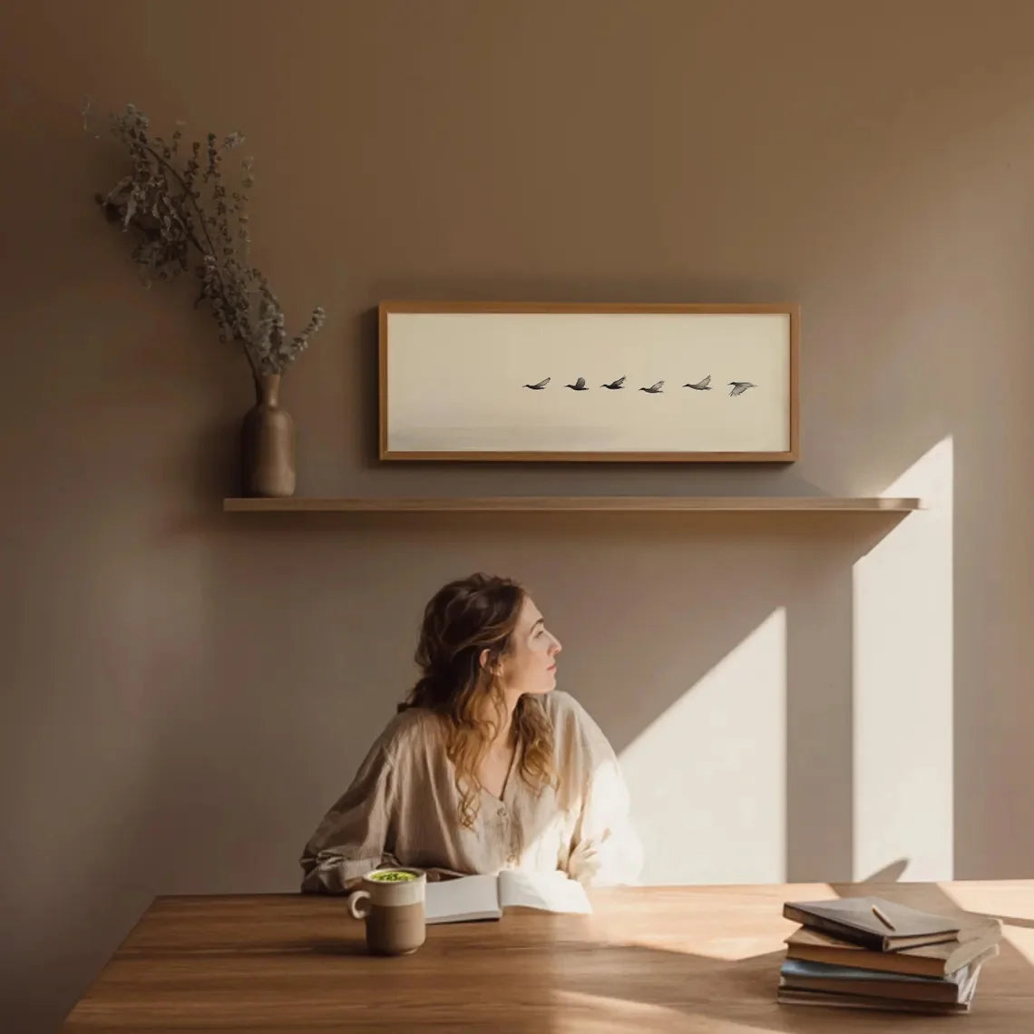 Woman sitting at a table with books and a mug, sunlit room with wall art.
