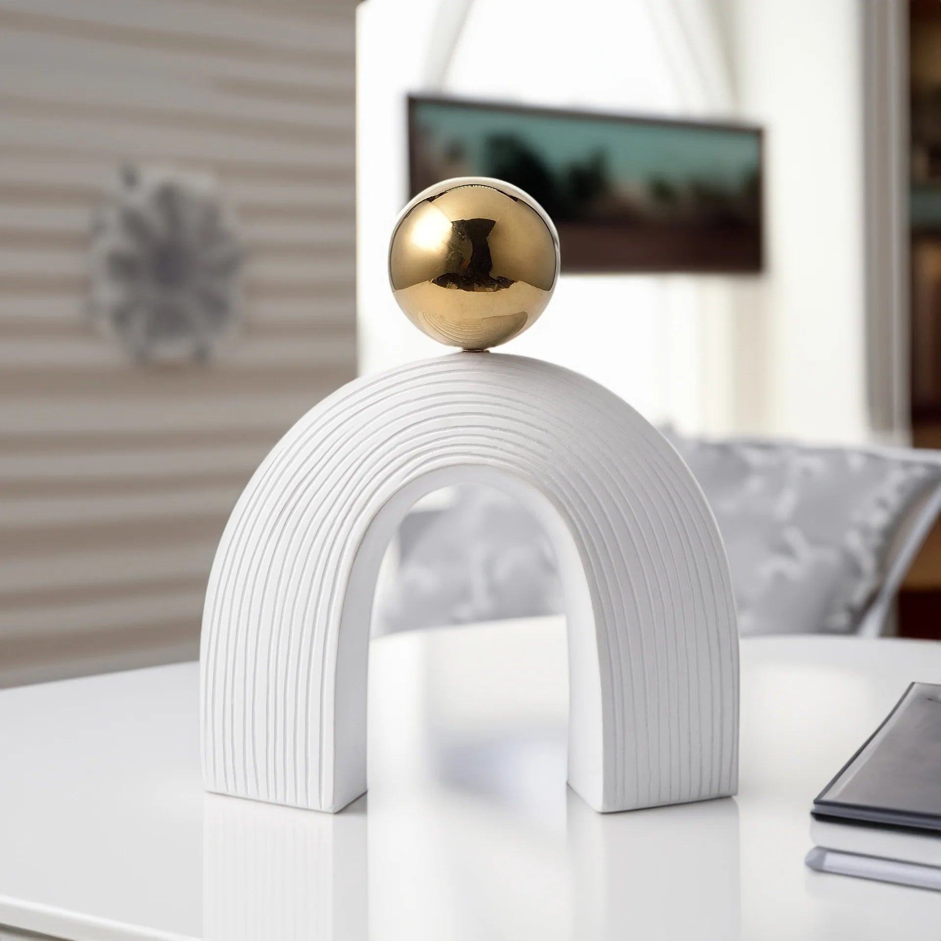 Decorative object with a gold sphere on a white arch-shaped base in a blurred indoor setting