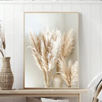 Framed artwork of pampas grass in a vase on a wooden surface with a white wall background.
