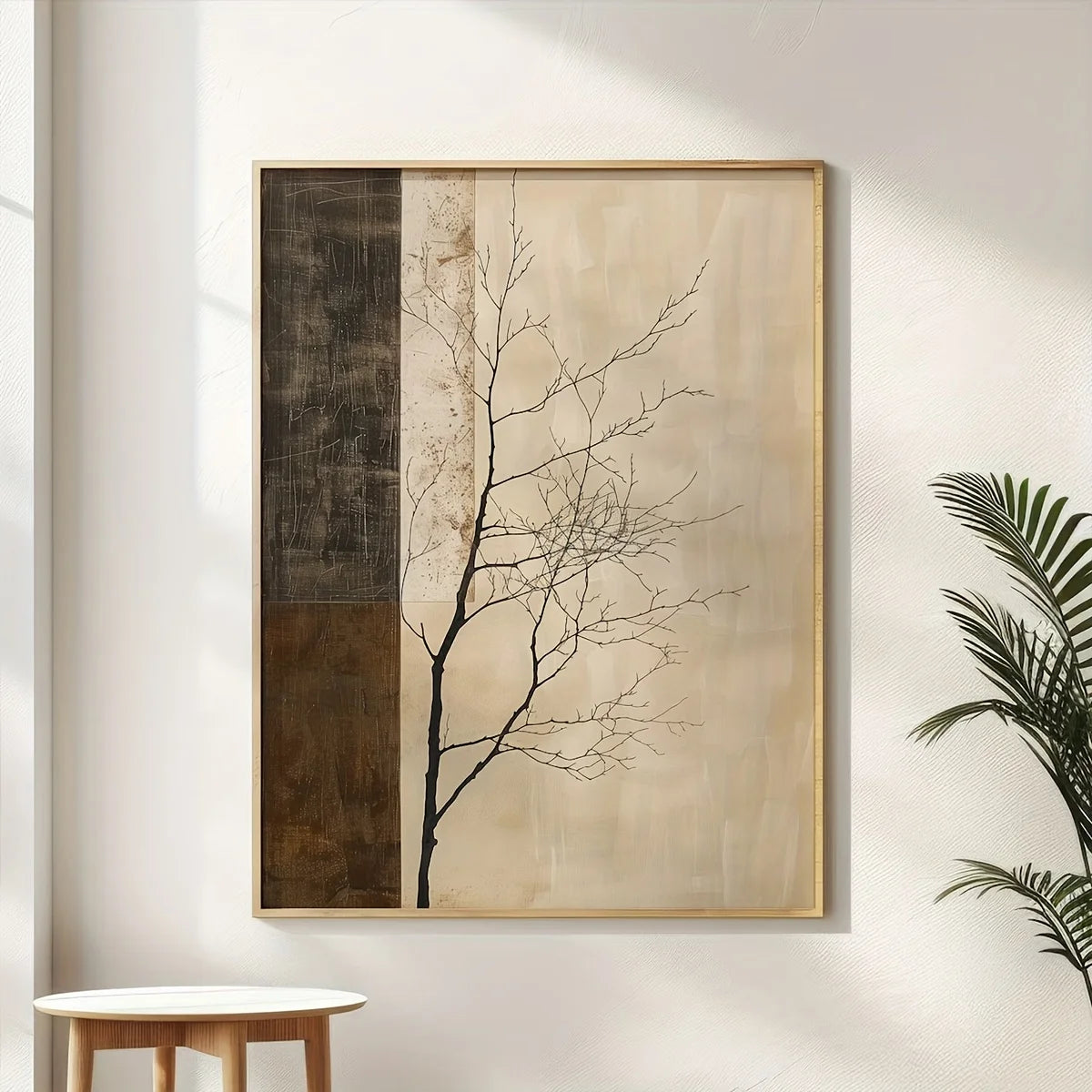 Framed wall art hanging on a white wall, featuring a minimalist black tree branch design over beige and dark brown textured panels, with a light wood frame. A small round table is partially visible at the bottom left, and a green plant is visible at the right edge.