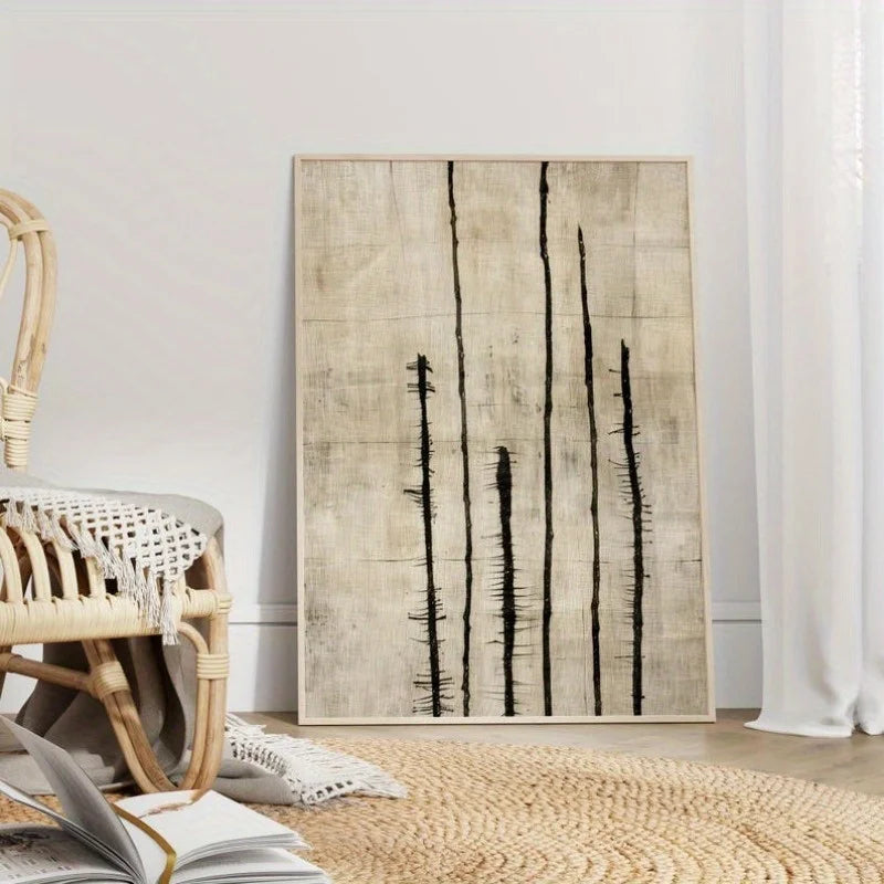 A vertical wall art print with a textured beige background and black vertical brushstroke lines, leaning against a white wall in a styled room with a woven chair, rug, and open book.