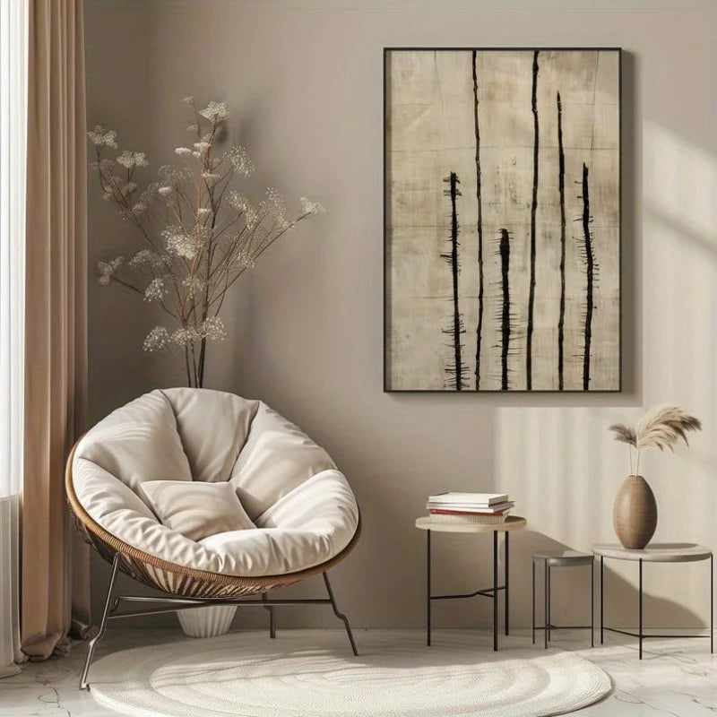 An abstract wall art print with vertical black brushstrokes on a textured beige background, displayed in a styled living room setting next to a cushioned wicker chair and side tables.