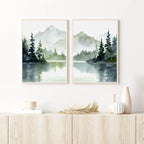 Two framed landscape paintings of mountains and trees over a lake on a white wall above a wooden shelf.
