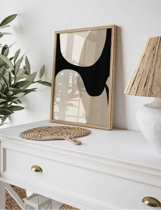 Framed abstract art on a white surface with a plant and lamp in the background