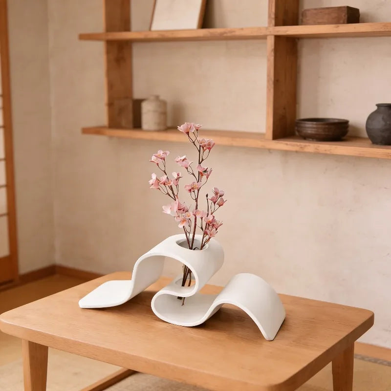 Modern white vase with pink flowers on a wooden table in a room with shelves.