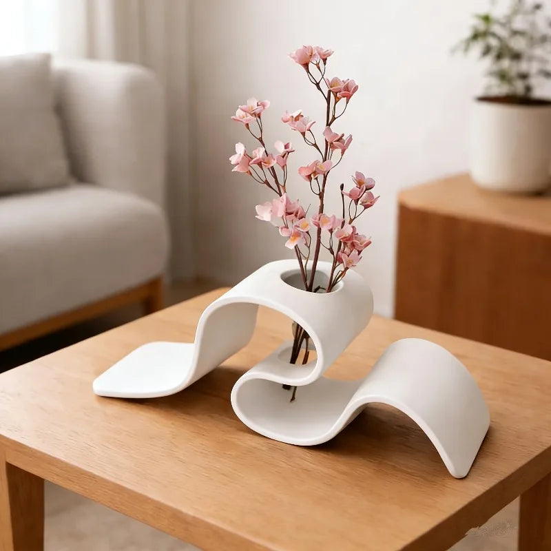 Modern white vase with pink flowers on a wooden table in a living room setting.