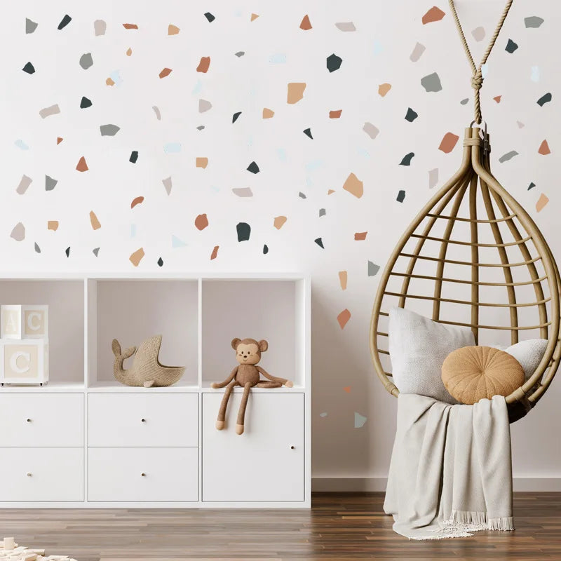 Children's room with a white bookshelf, teddy bear, hanging rattan chair, and colorful wall pattern.