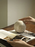Person holding a spherical concrete object over an open book on a wooden surface.