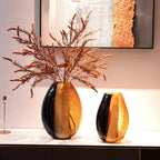 Two decorative vases with a black and gold finish on a surface, with a abstract painting in the background.