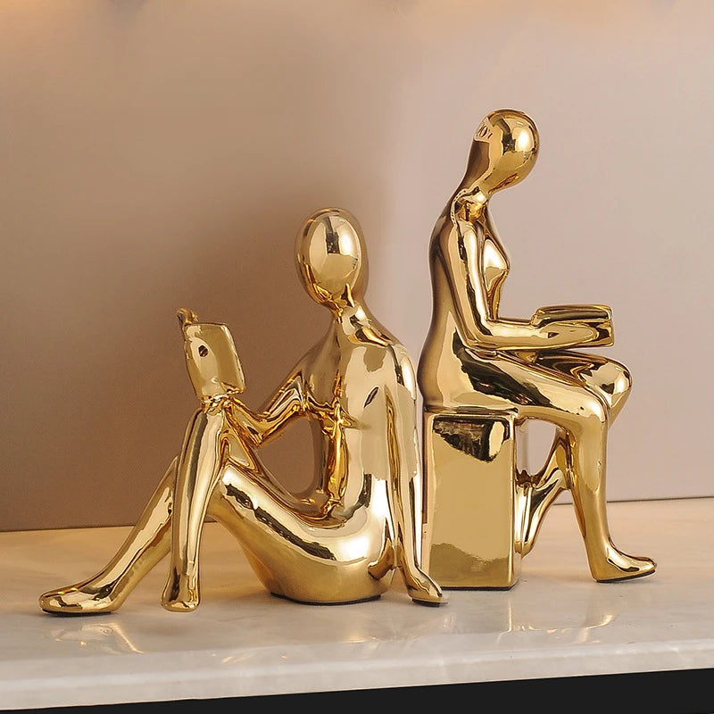 Two gold-colored sculptures of people sitting on a white surface with a beige background