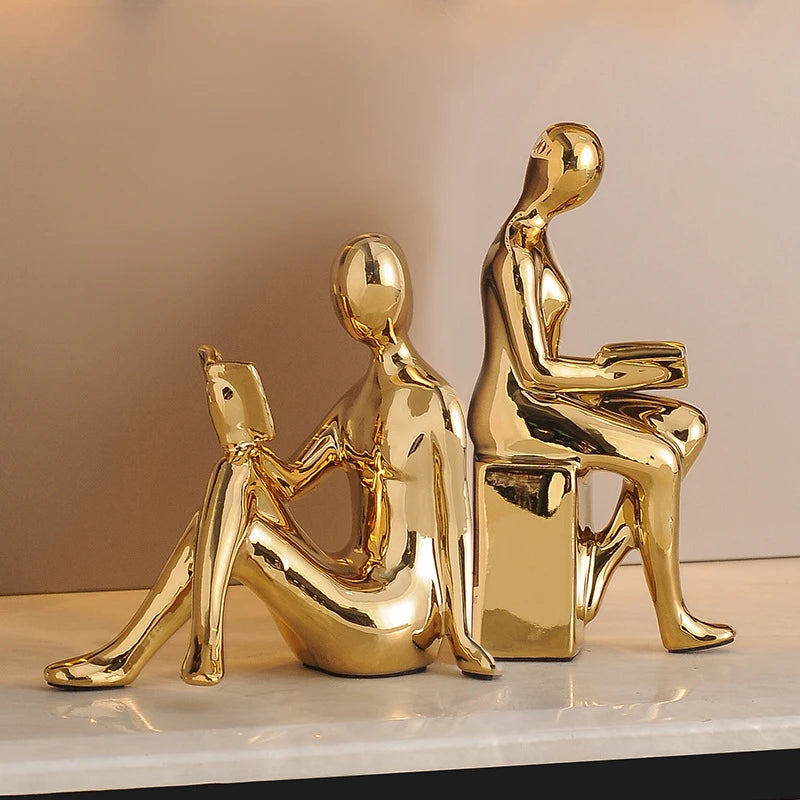 Two gold-colored sculptures of people sitting on a white surface with a beige background