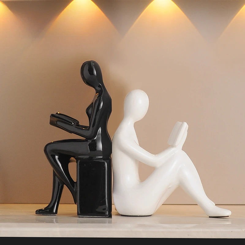 Two abstract sculptures, one black and one white, on a wooden surface with a beige wall background.