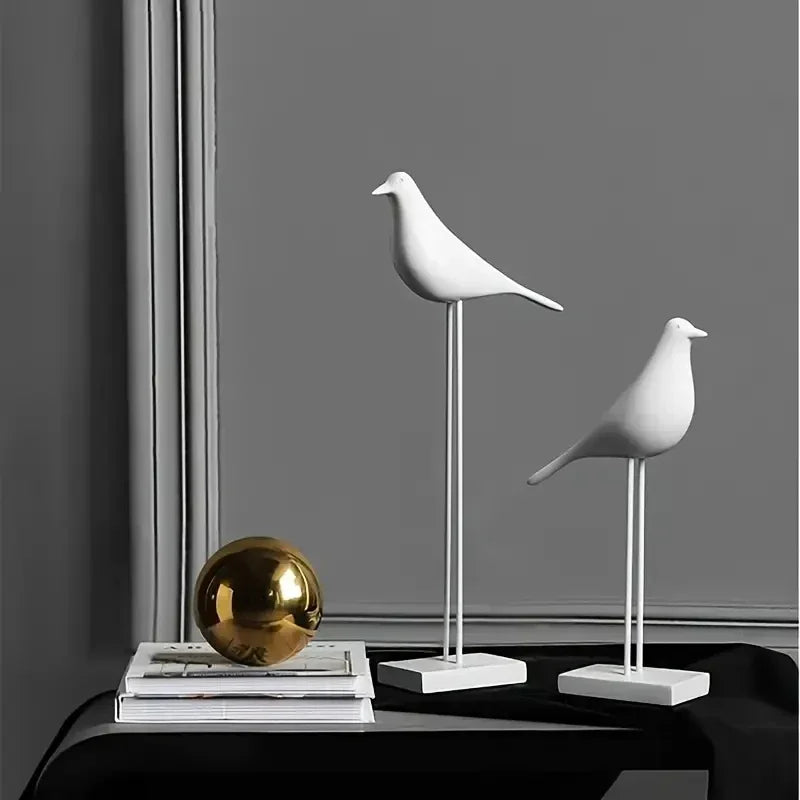 Two white bird sculptures on stands with a gold sphere and books on a dark surface against a gray wall.