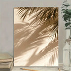 Framed artwork of palm leaves casting shadows on a wall with books and a plant in the foreground.