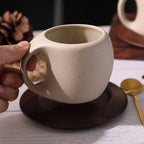Beige ceramic mug with handle held by a hand on a wooden surface.