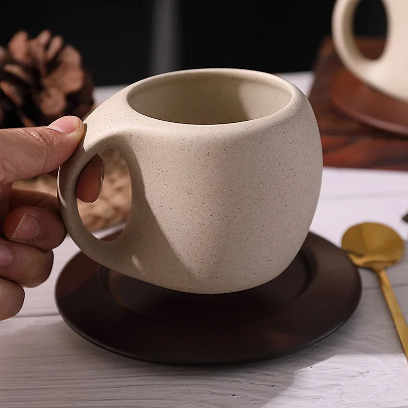 Beige ceramic mug with handle held by a hand on a wooden surface.
