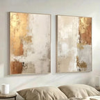 Two abstract art pieces in gold and beige tones on a wall above a bed.