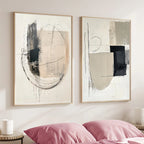 Two abstract art pieces on a wall above a bed with pink pillows.