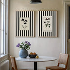 Two botanical art prints with black and white vertical stripes hang on a beige wall above a round dining table with chairs, a blue vase of purple flowers, and a bowl of fruit.